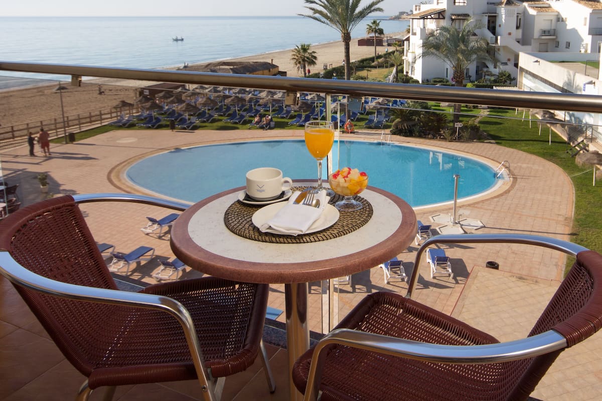 A poolside table with a drink, a bowl of fruit, and a cup, overlooking a beach and a pool with lounge chairs and umbrellas.