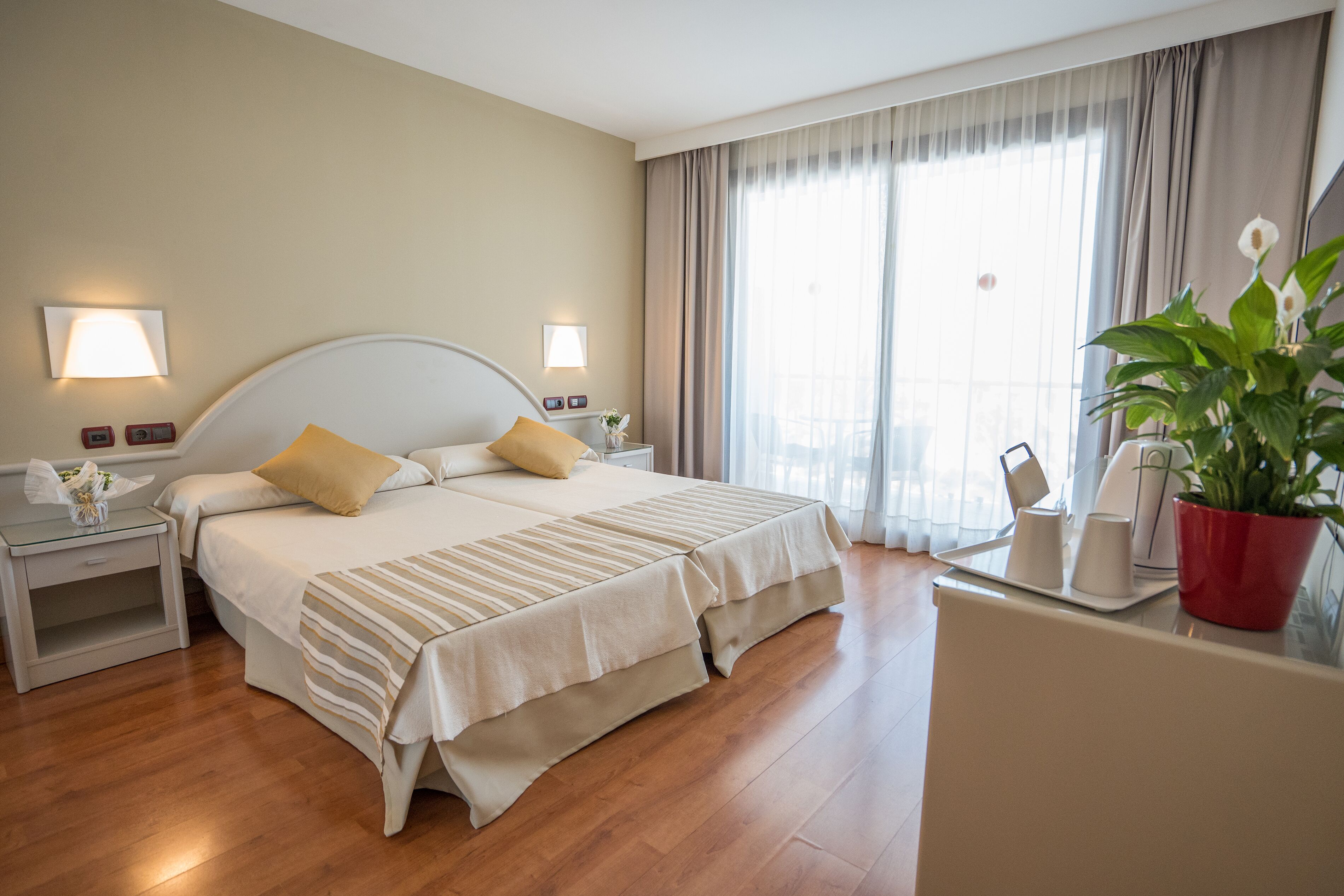 A hotel room with a large bed, bedside tables, a desk with a chair, and a potted plant.