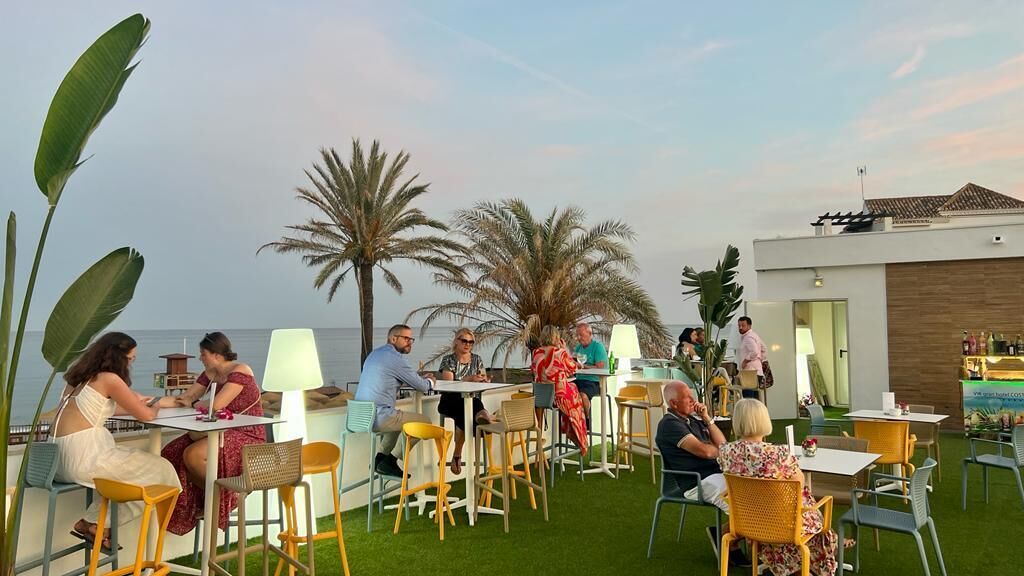 An outdoor dining area with tables and chairs, palm trees, and a view of the sea.
