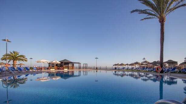 Una piscina azul cristalina con sillas de descanso y sombrillas, rodeada de palmeras y una zona de estar cubierta.
