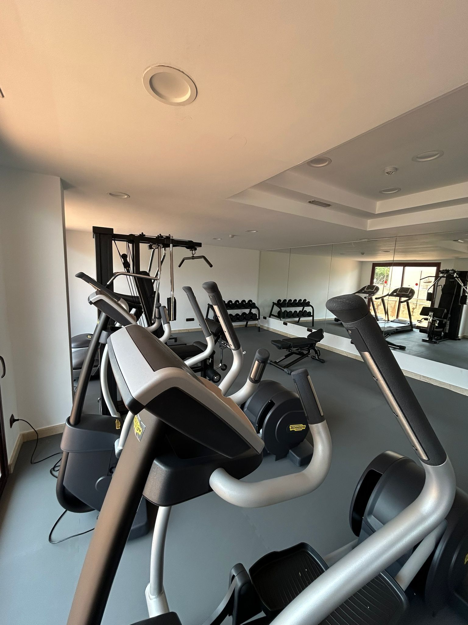 A modern gym with multiple exercise machines, including treadmills and stationary bikes, and a large mirror wall.