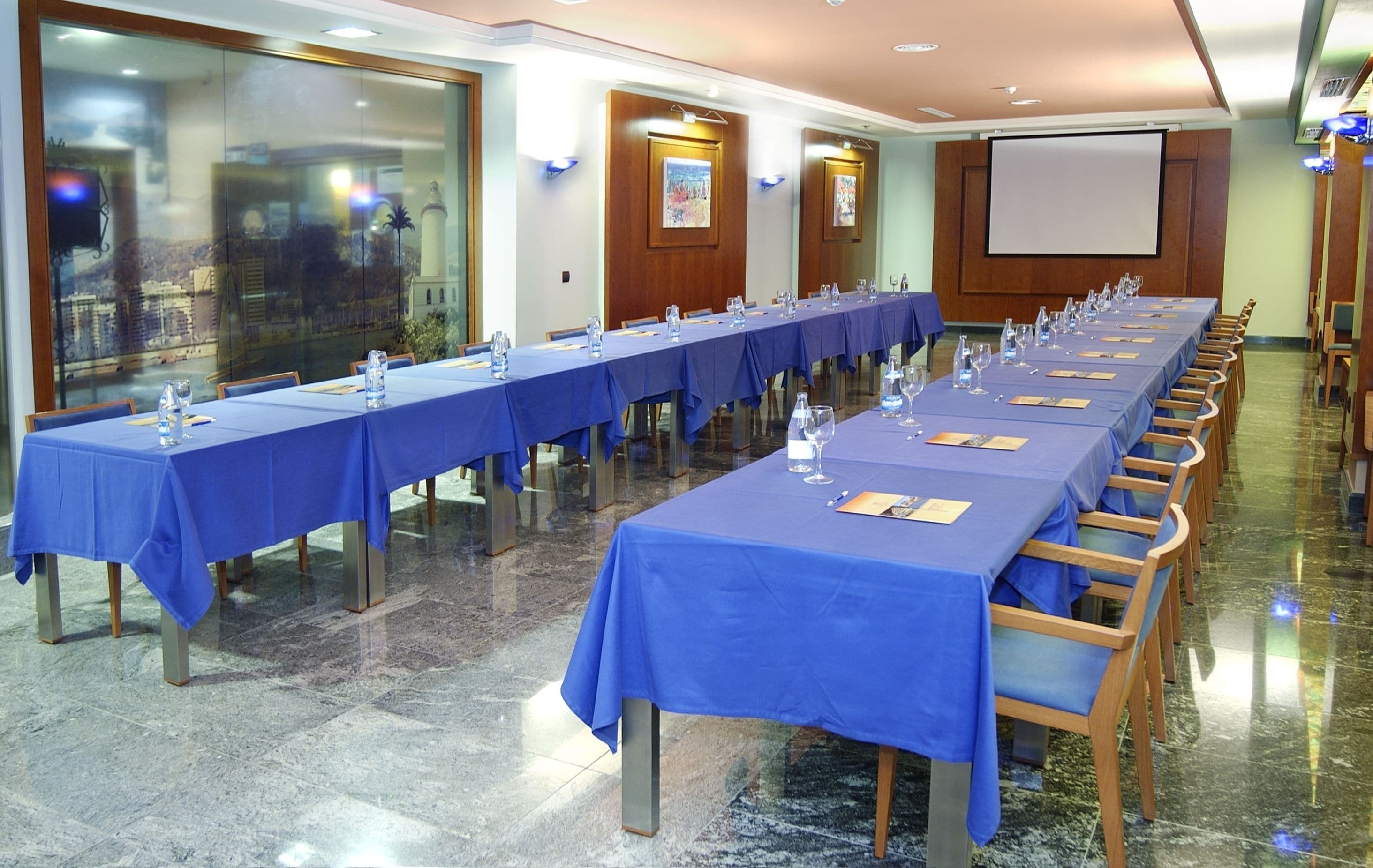 A conference room with long tables covered in blue tablecloths, equipped with water glasses, wine glasses, and notepads.