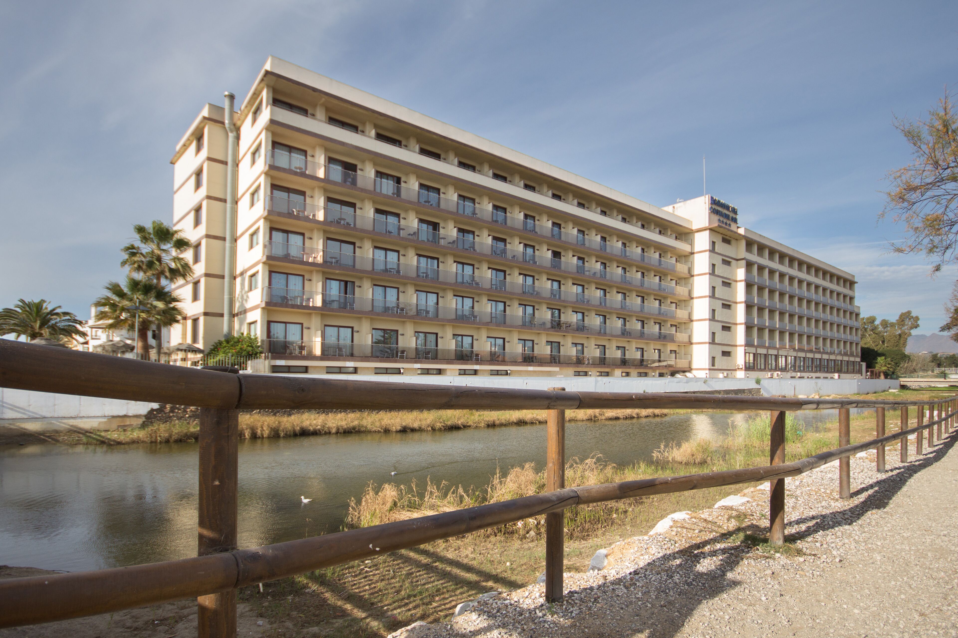 A multi-story hotel with balconies, a river in front, and a wooden fence.
