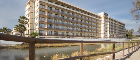 A multi-story hotel with balconies, a river in front, and a wooden fence.