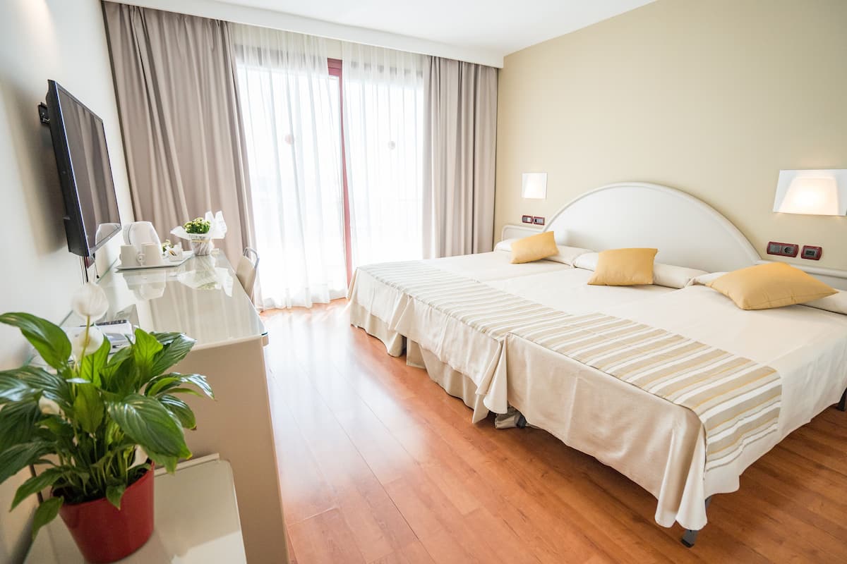A hotel room with a large bed, bedside lamps, a flat-screen TV, a desk with a plant, and a window with curtains.