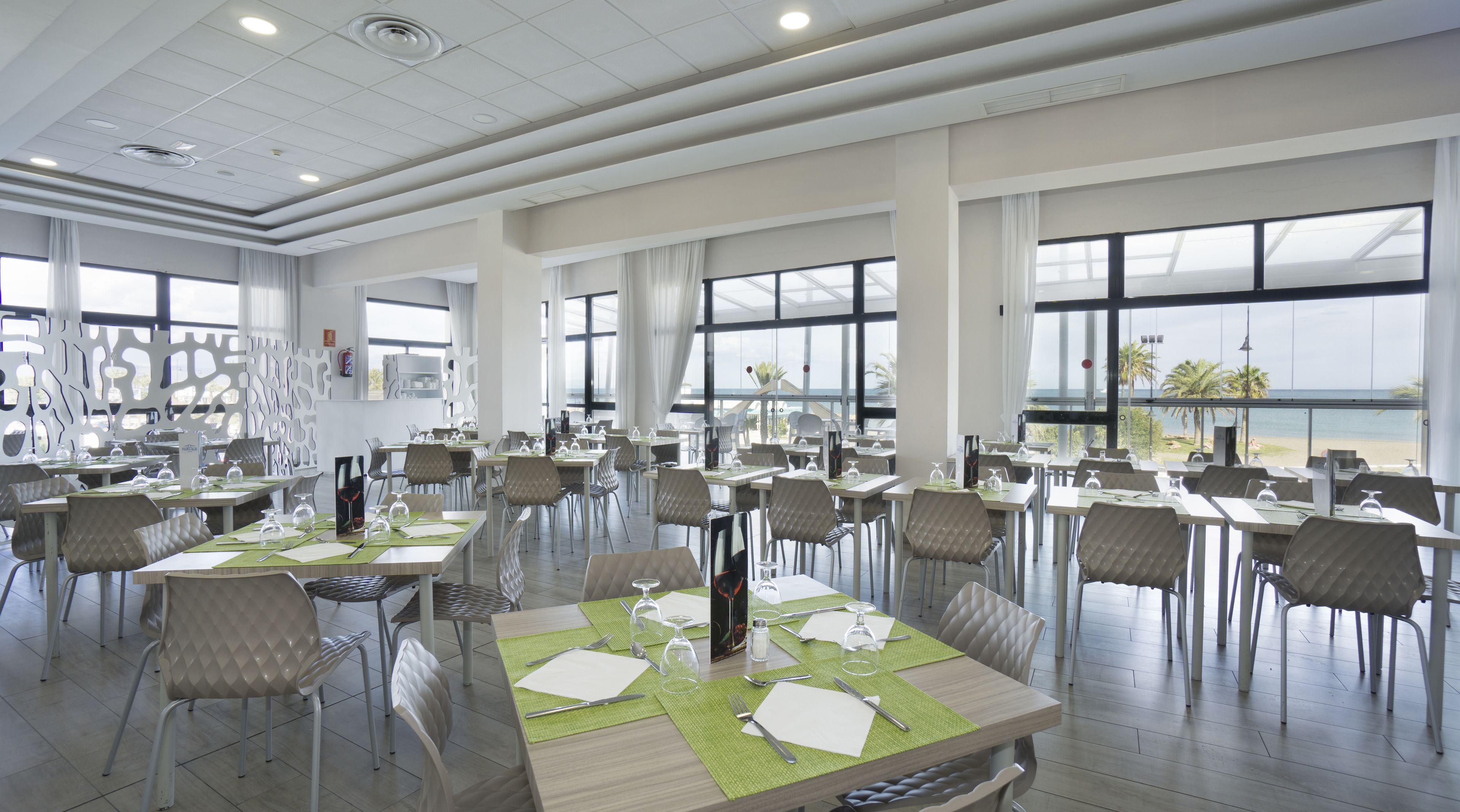 Un vaste espace dédié à la salle à manger, avec de nombreuses tables et chaises, de grandes fenêtres offrant une vue sur la plage et un design moderne au niveau du plafond.