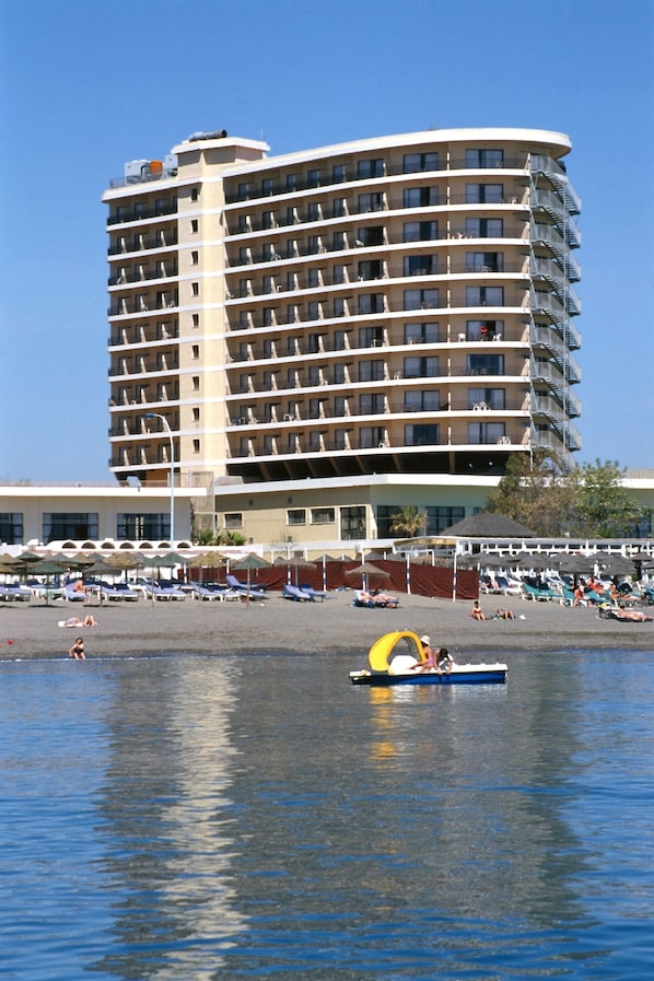Un hotel de varios pisos con balcones, una zona de playa con tumbonas y un barco en el agua.