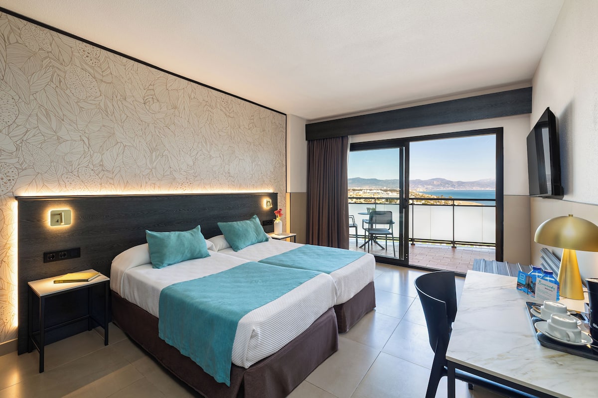A hotel room with a bed, bedside table, lamp, and a view of the sea through a balcony.