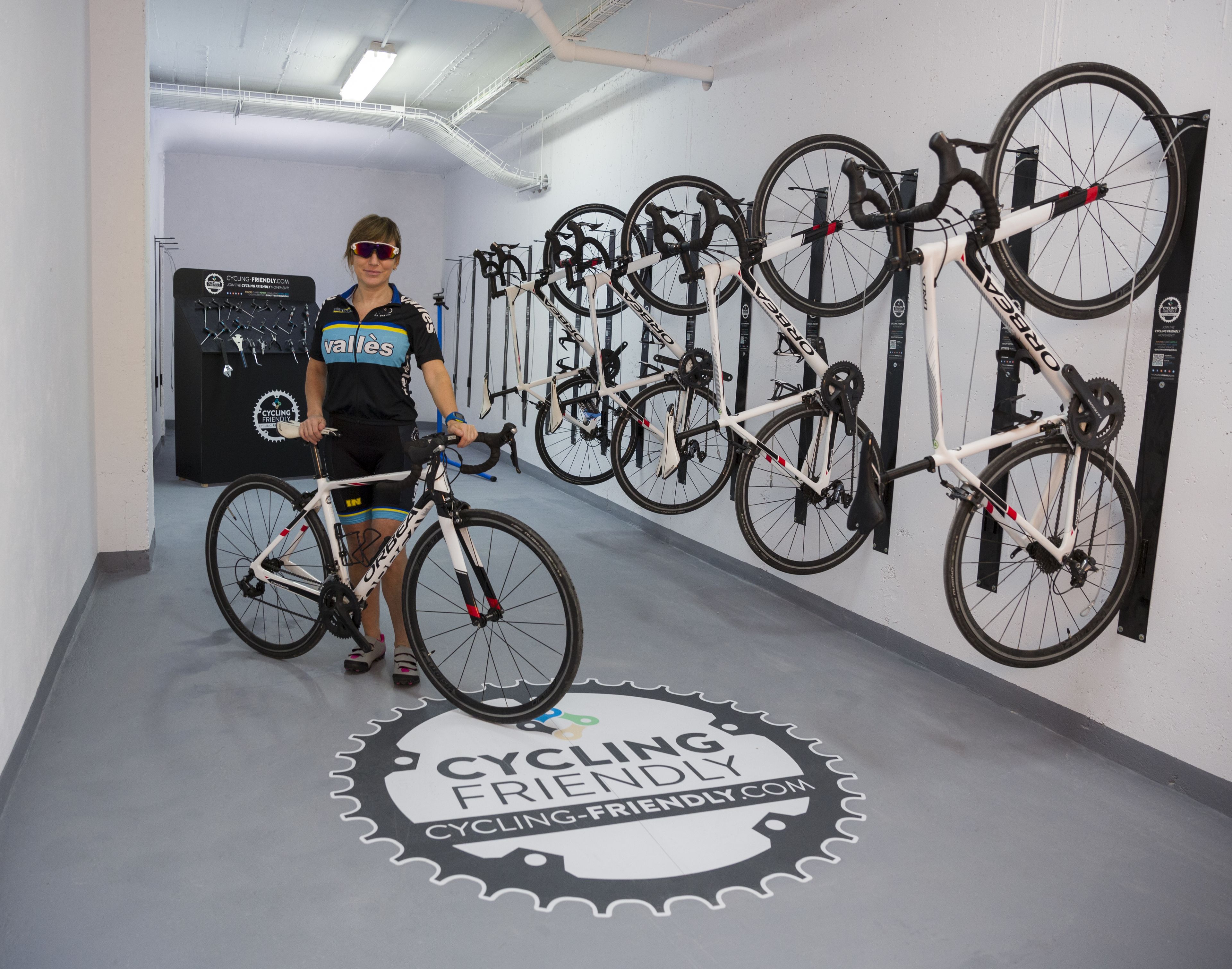 Un local de alquiler de bicicletas con varias bicicletas colgadas en estantes de pared, un logotipo en el suelo y una persona de pie junto a una bicicleta.