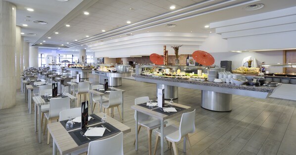 Un comedor moderno con varias mesas y sillas, una barra de buffet con distintos alimentos, y un ambiente limpio y bien iluminado.