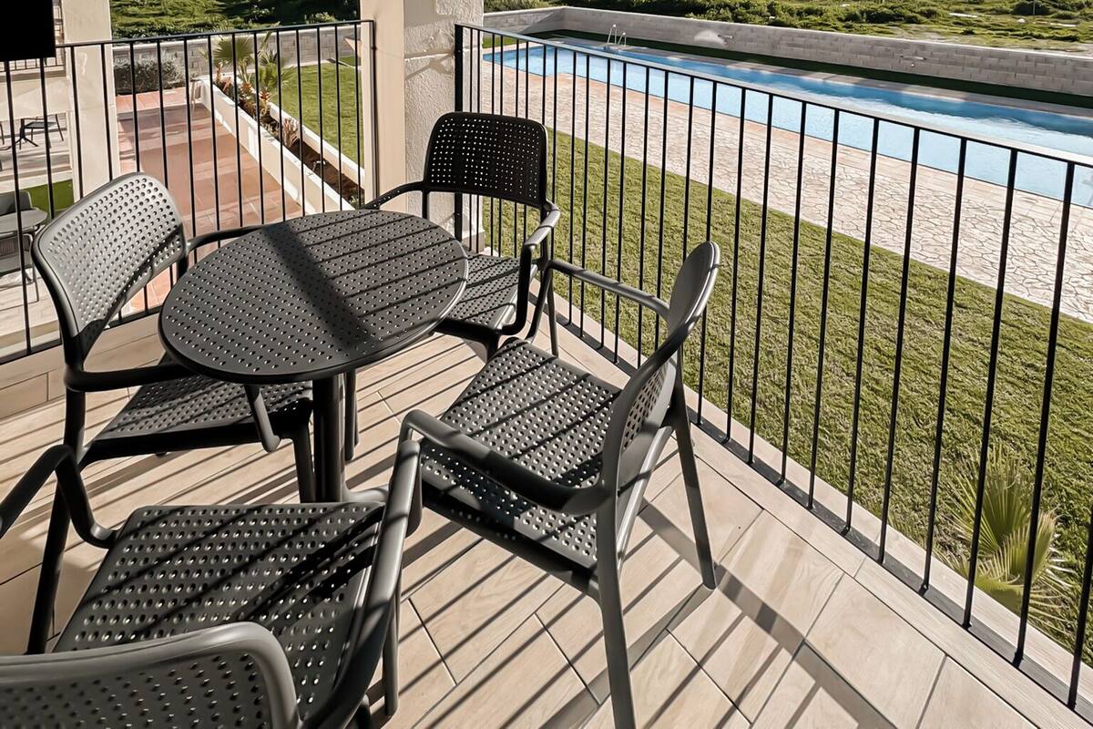 Un balcón con vista a la piscina, mobiliario exterior y edificios al fondo.