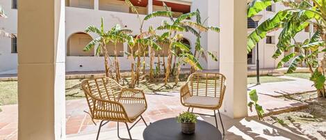 An outdoor patio with wicker chairs and a small table, surrounded by banana trees and a white building with balconies.