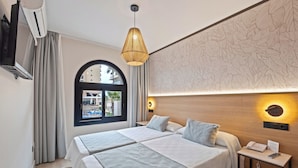 A hotel room with two beds, a TV, a window with a view, and a bedside table with a phone and a lamp. - Hotel Puente Real (Torremolinos)