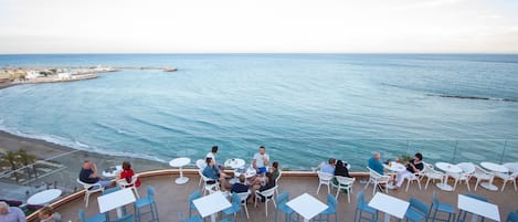 A terrace with tables and chairs overlooking the sea, with a view of the coastline and a pier in the distance.