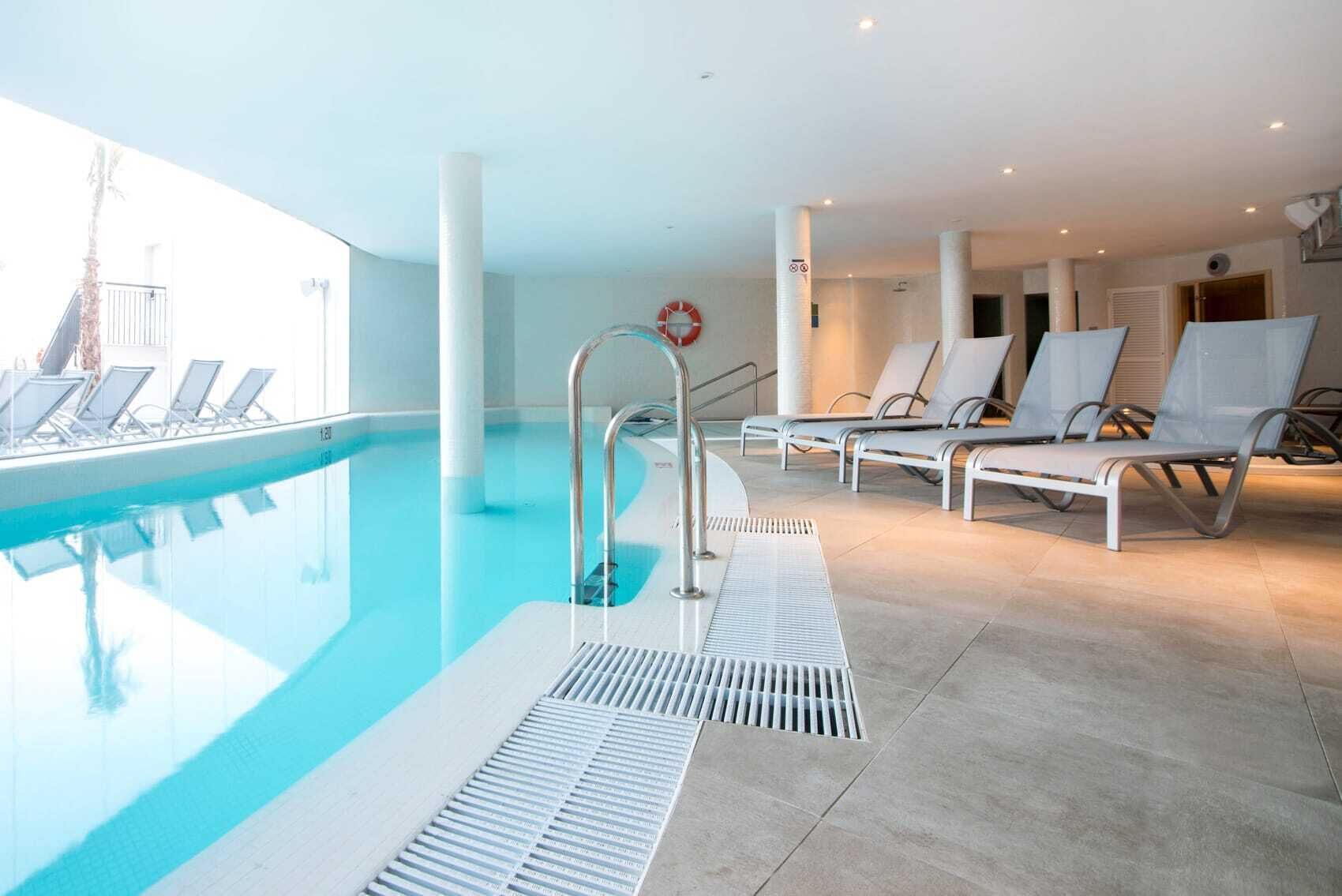 Una piscina interior con agua cristalina, una escalera metálica y varias sillas reclinables.