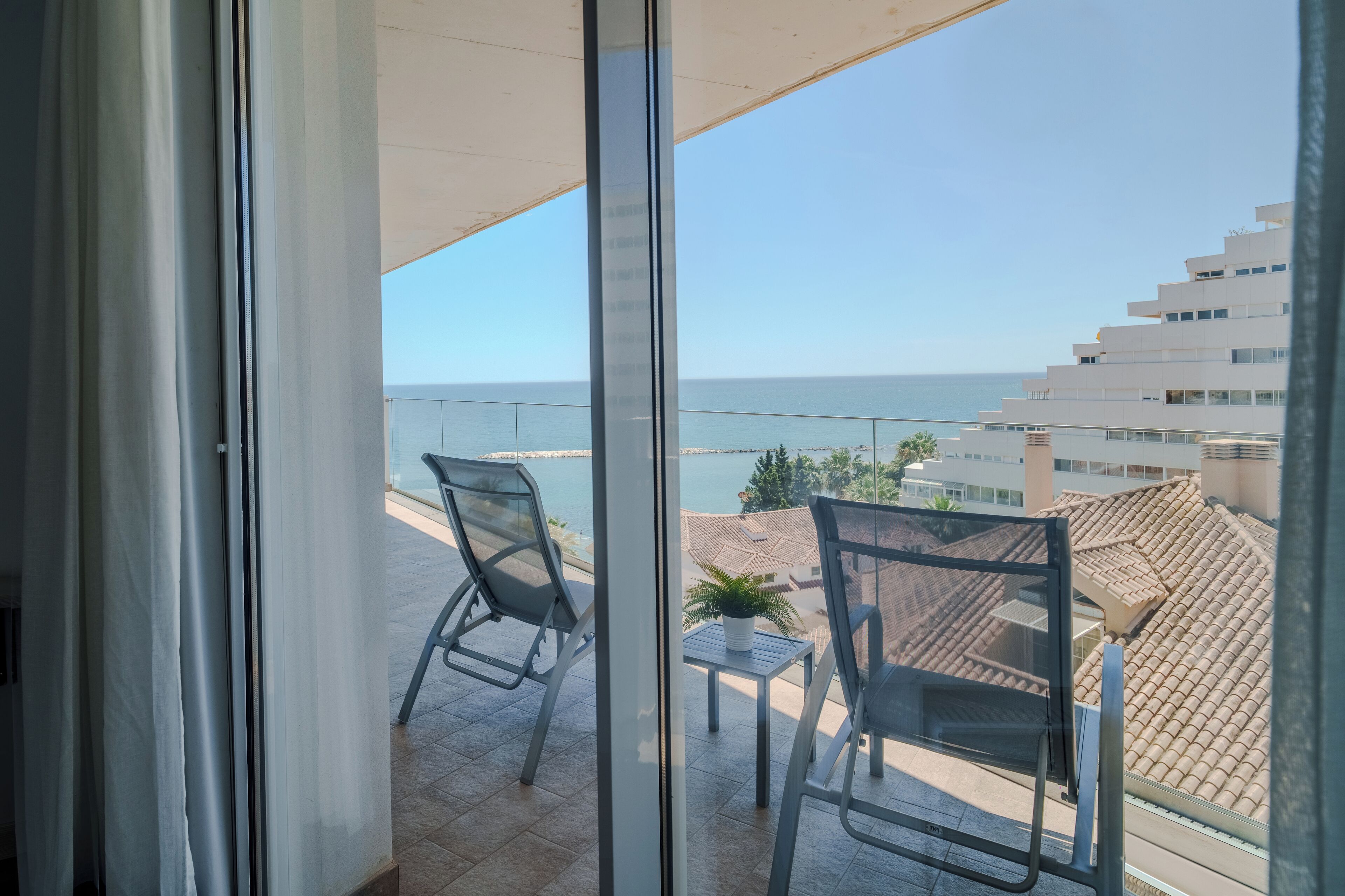 Un balcón con dos sillas y una mesita, con vistas a la costa y edificios al fondo.
