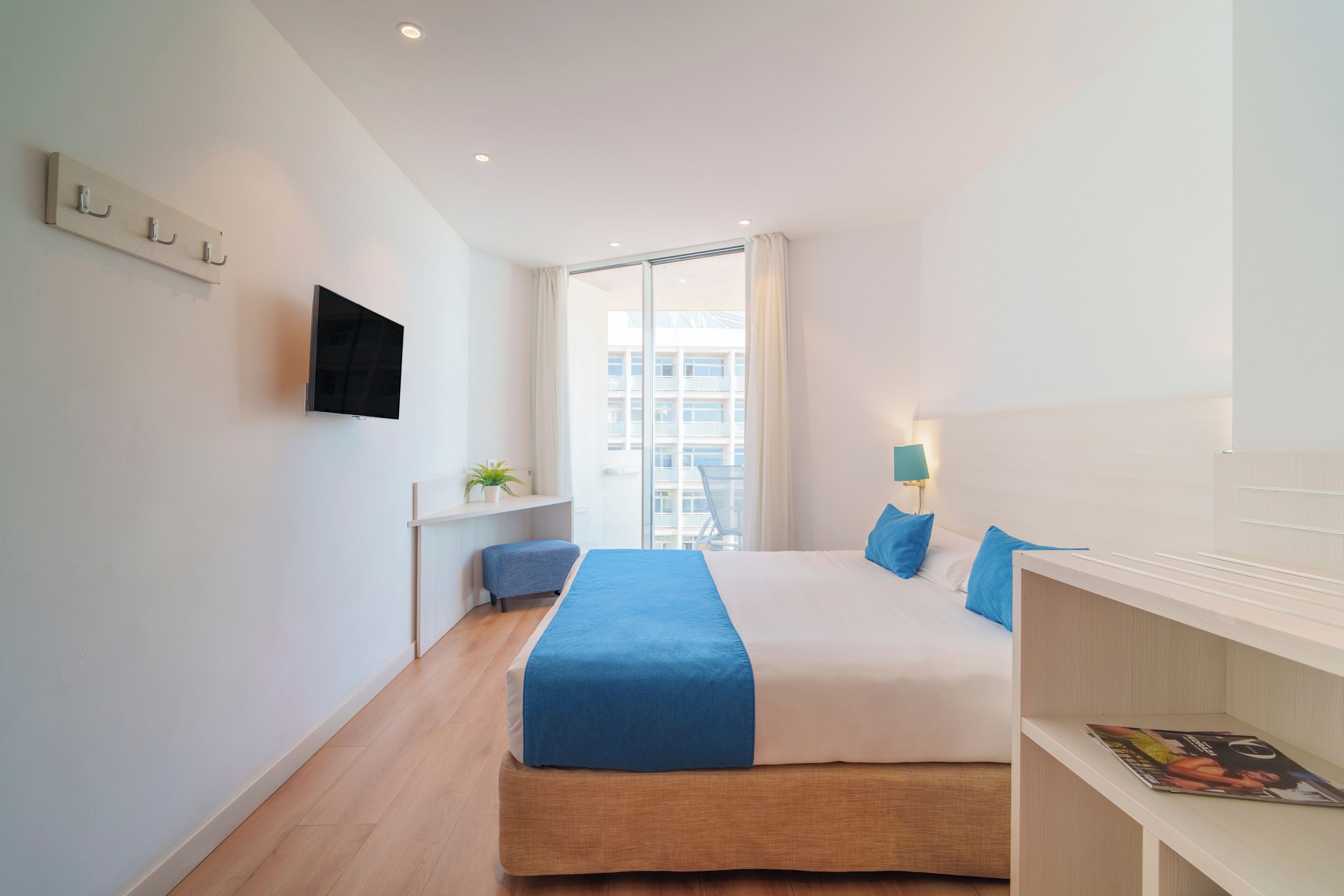 Una habitación de hotel moderna con una cama grande, un televisor de pantalla plana montado en la pared, un pequeño rincón de descanso con una planta y una ventana con vistas a la ciudad.