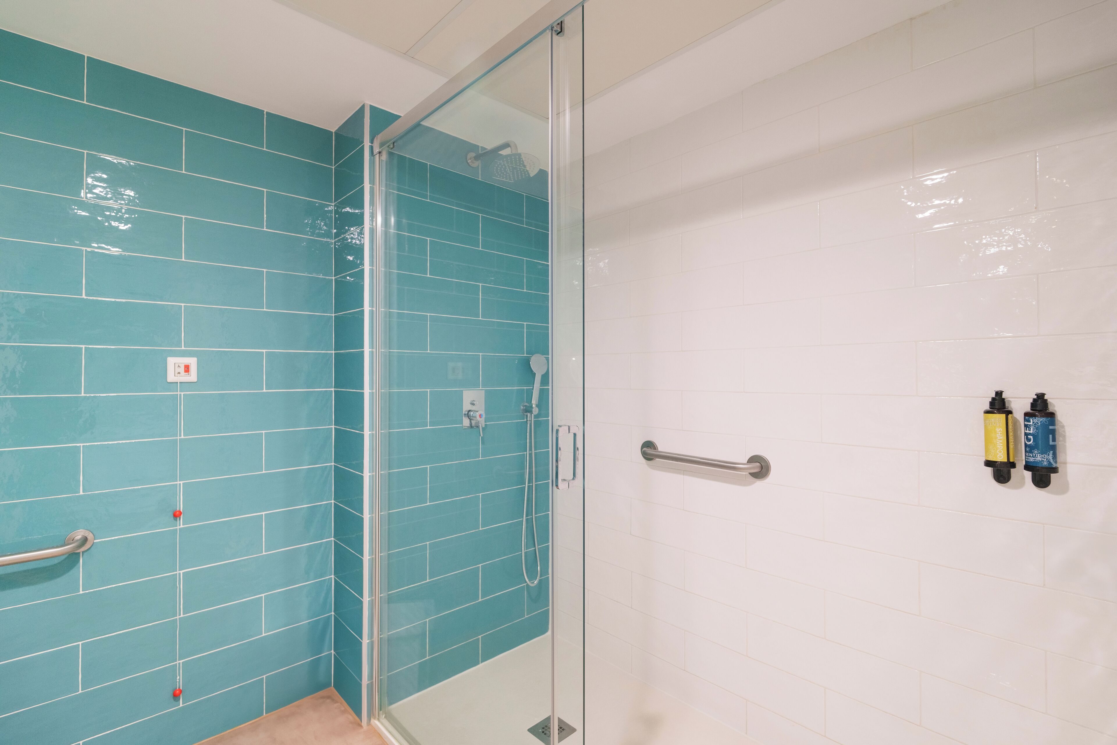 Un baño con azulejos verde mar y blancos, cerramiento de ducha de vidrio y barras de apoyo.