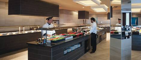 Disposição moderna de buffet com chefs preparando alimentos, apresentando uma ilha central com diversos itens e um balcão extenso com displays de comida.