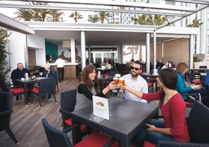 An outdoor dining area with wicker chairs and red cushions, a menu stand, and a view of the interior through glass doors.
