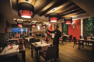 Un restaurant avec plusieurs tables et chaises, un serveur qui s’occupe d’une table, et une vue sur la mer par les fenêtres.