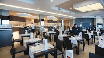 Un restaurante moderno con mesas dispuestas para comer, un chef preparando comida y una zona de bar al fondo.