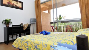 A hotel room with a bed, a desk with a chair, a television, a window with curtains, and a balcony with chairs and plants.