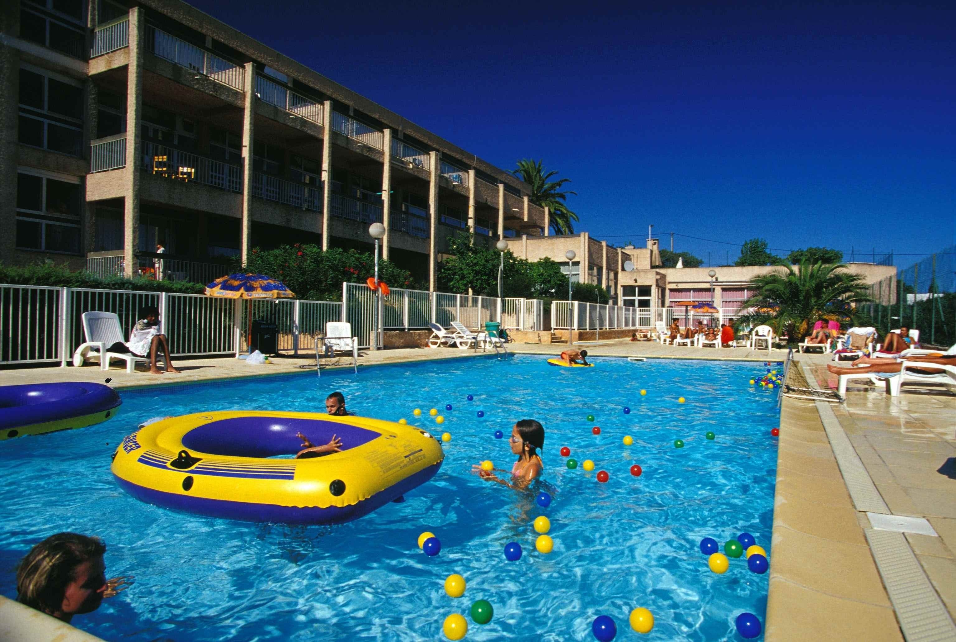 A swimming pool with inflatable rings and colorful balls, surrounded by lounge chairs and a building with balconies.