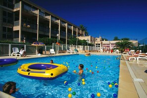 A swimming pool with inflatable rings and colorful balls, surrounded by lounge chairs and a building with balconies.