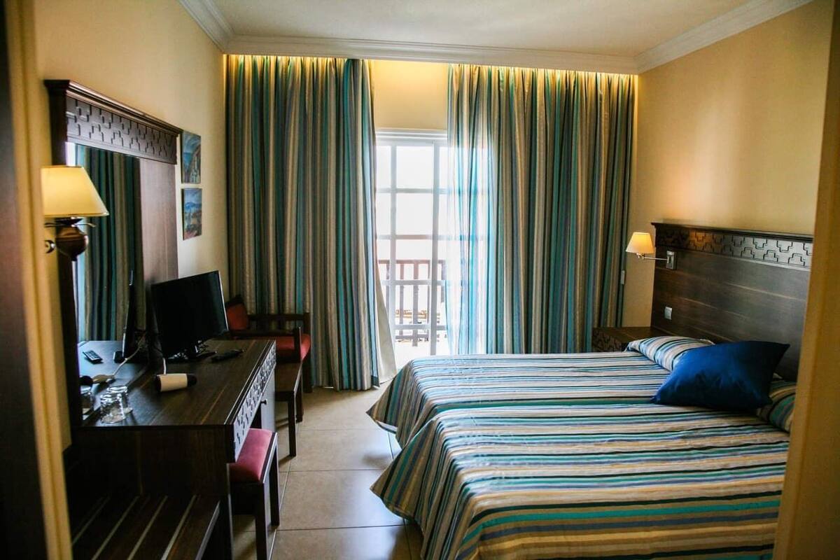 Una habitación de hotel con cama, escritorio, silla y un ventanal con cortinas.