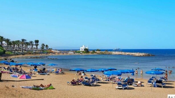 A beach with numerous sun loungers and umbrellas, a white building in the background, and a calm sea. - Anais Bay Hotel (Paralimni)