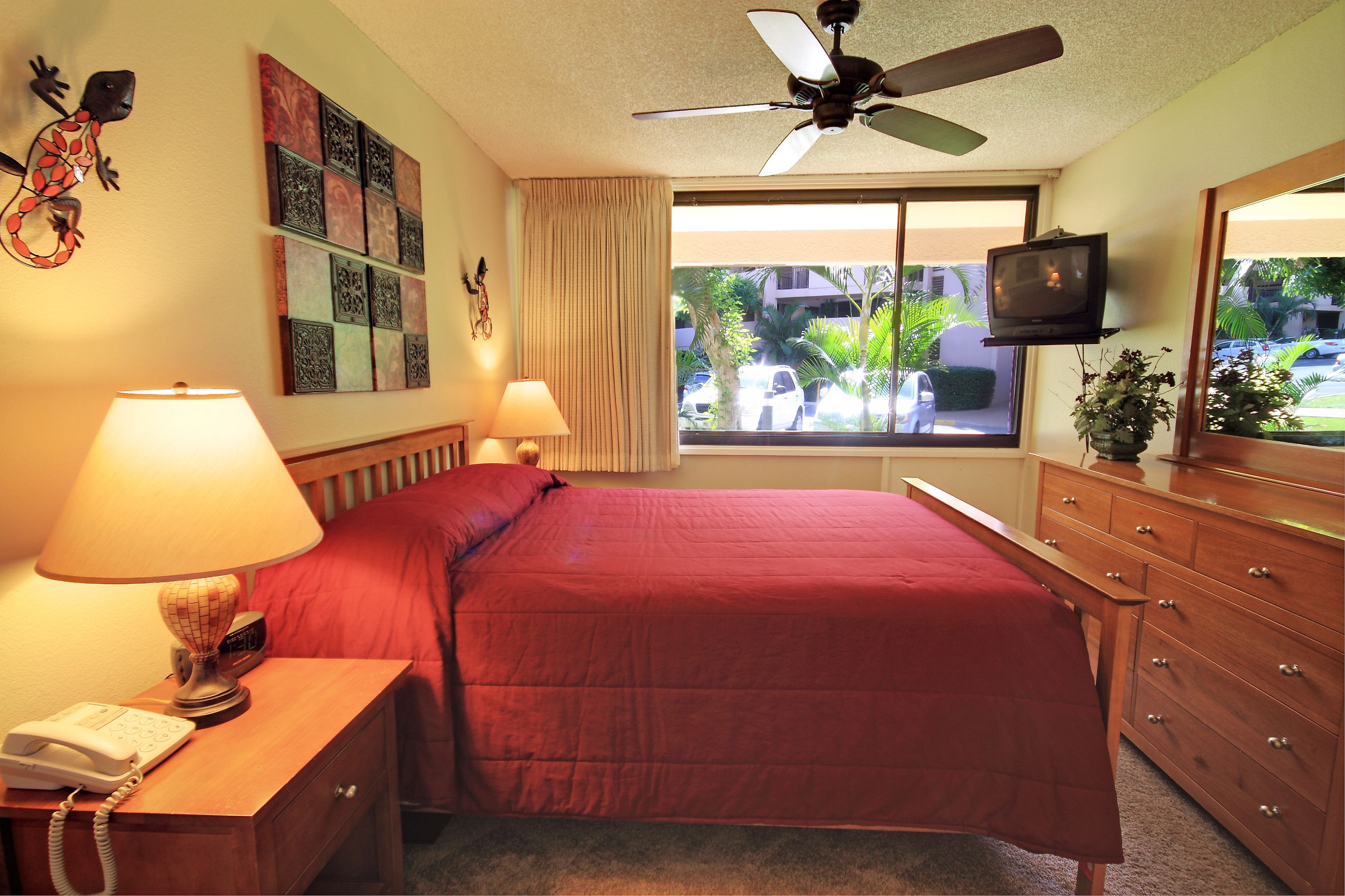A hotel room with a bed, a nightstand with a lamp, a dresser, a ceiling fan, a TV, and a window with a view of the outside.