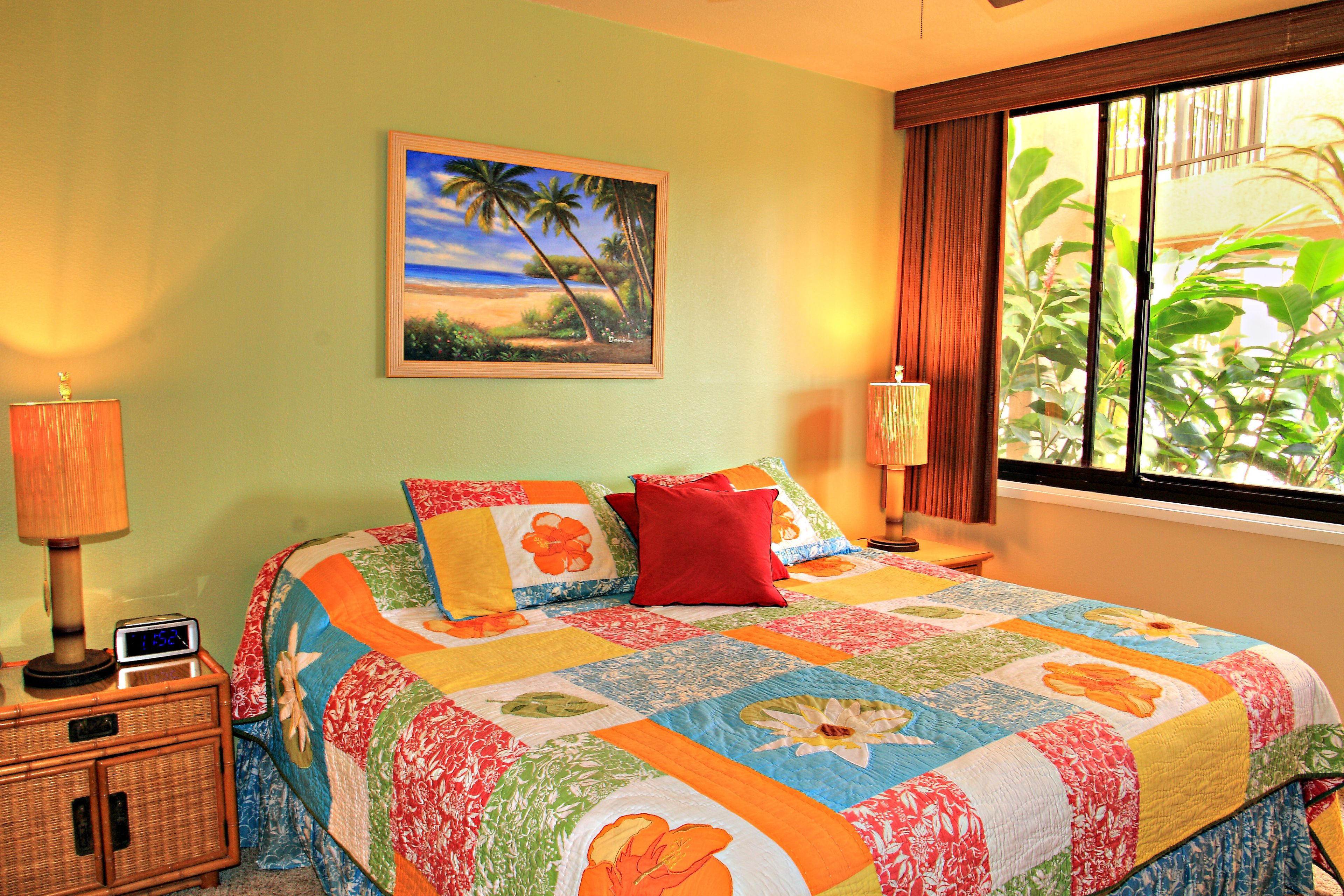 Una cama con un colcha de colores, una mesita de noche con una lámpara, un cuadro de una playa y una ventana con vistas a la vegetación.