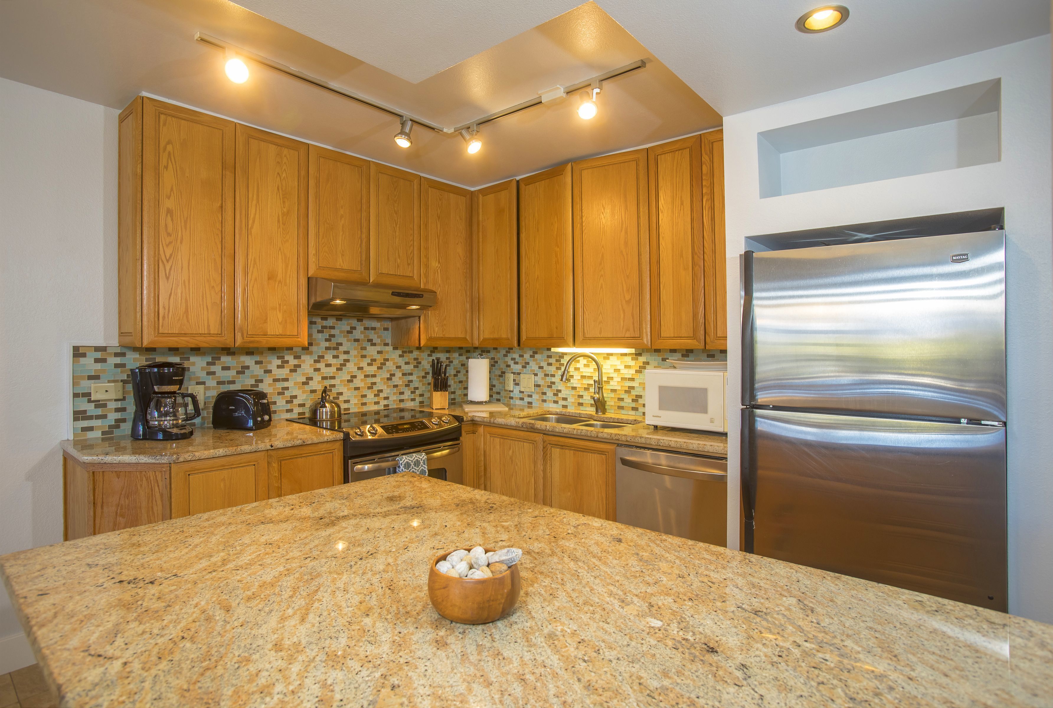 A kitchen with wooden cabinets, a stainless steel refrigerator, granite countertops, and a tiled backsplash.