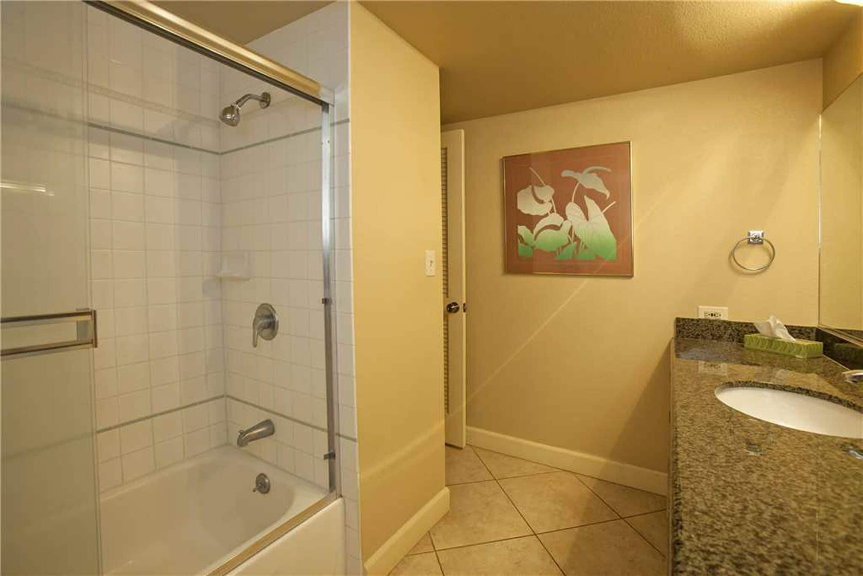 Un baño con ducha envuelta en vidrio, un lavamanos con encimera de granito y un aro para toallas en la pared.