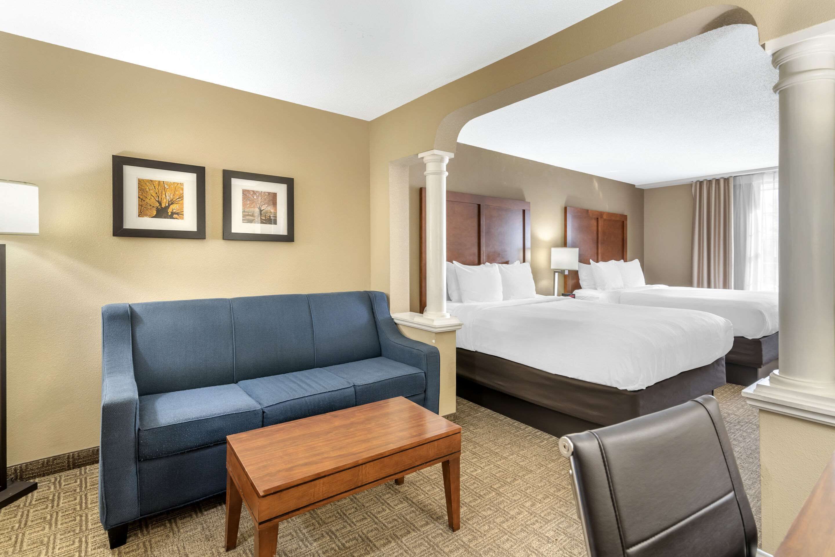 Un cuarto de hotel con una cama grande, un sofá azul, una mesa de centro de madera y dos cuadros en la pared.