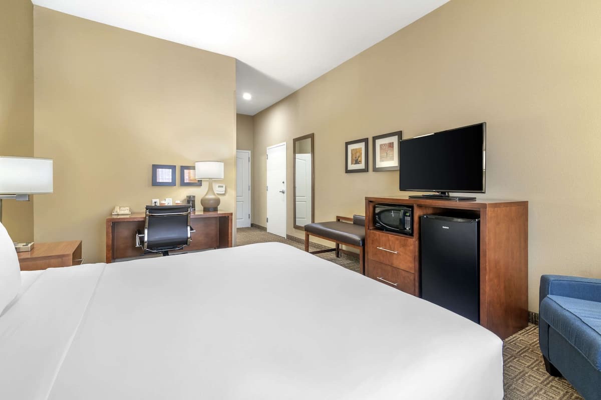 A hotel room with a large bed, a desk with a chair, a television, a refrigerator, and a blue armchair.