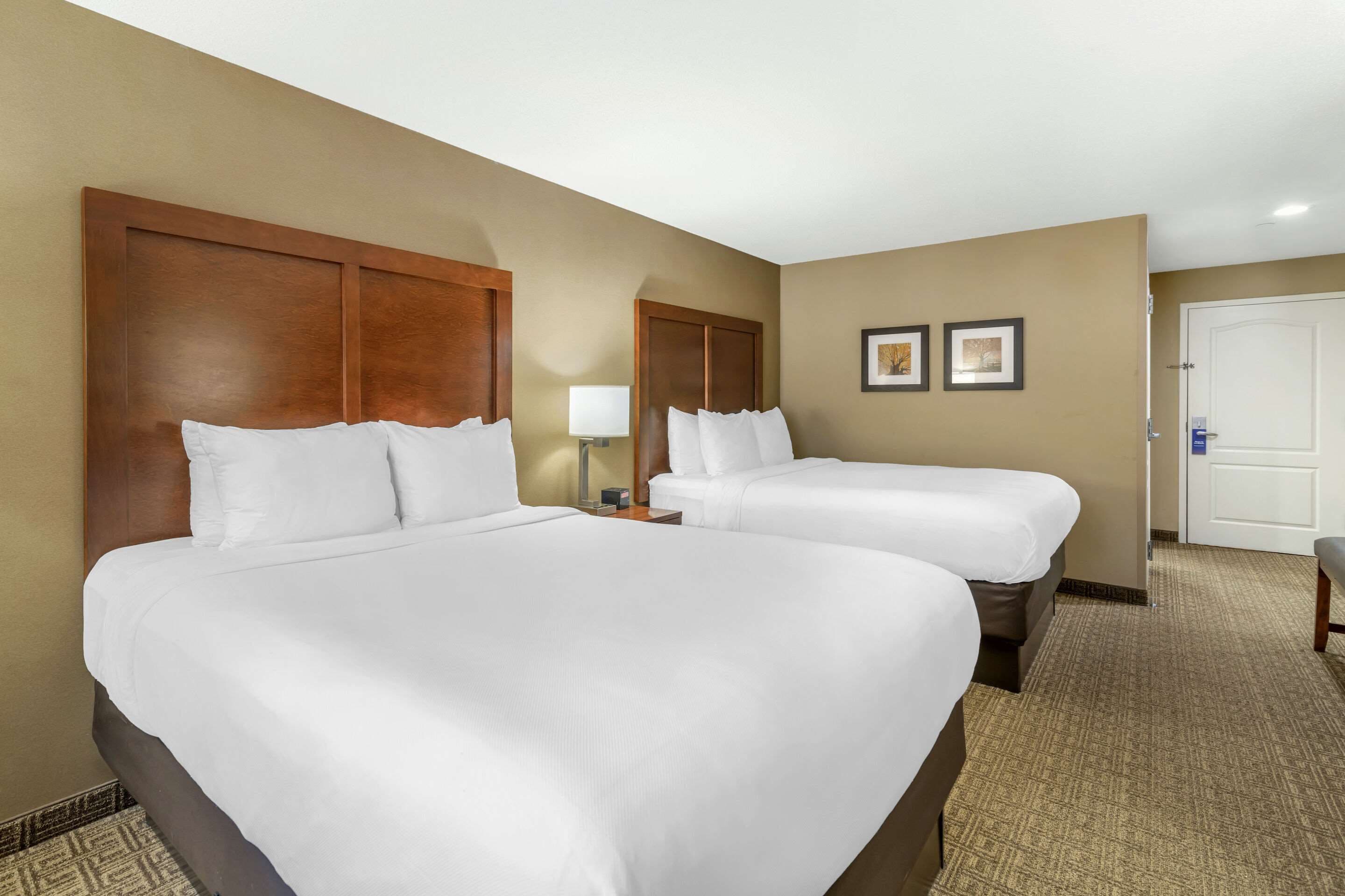 A hotel room with two large beds, a wooden headboard, a nightstand, a lamp, and a door.