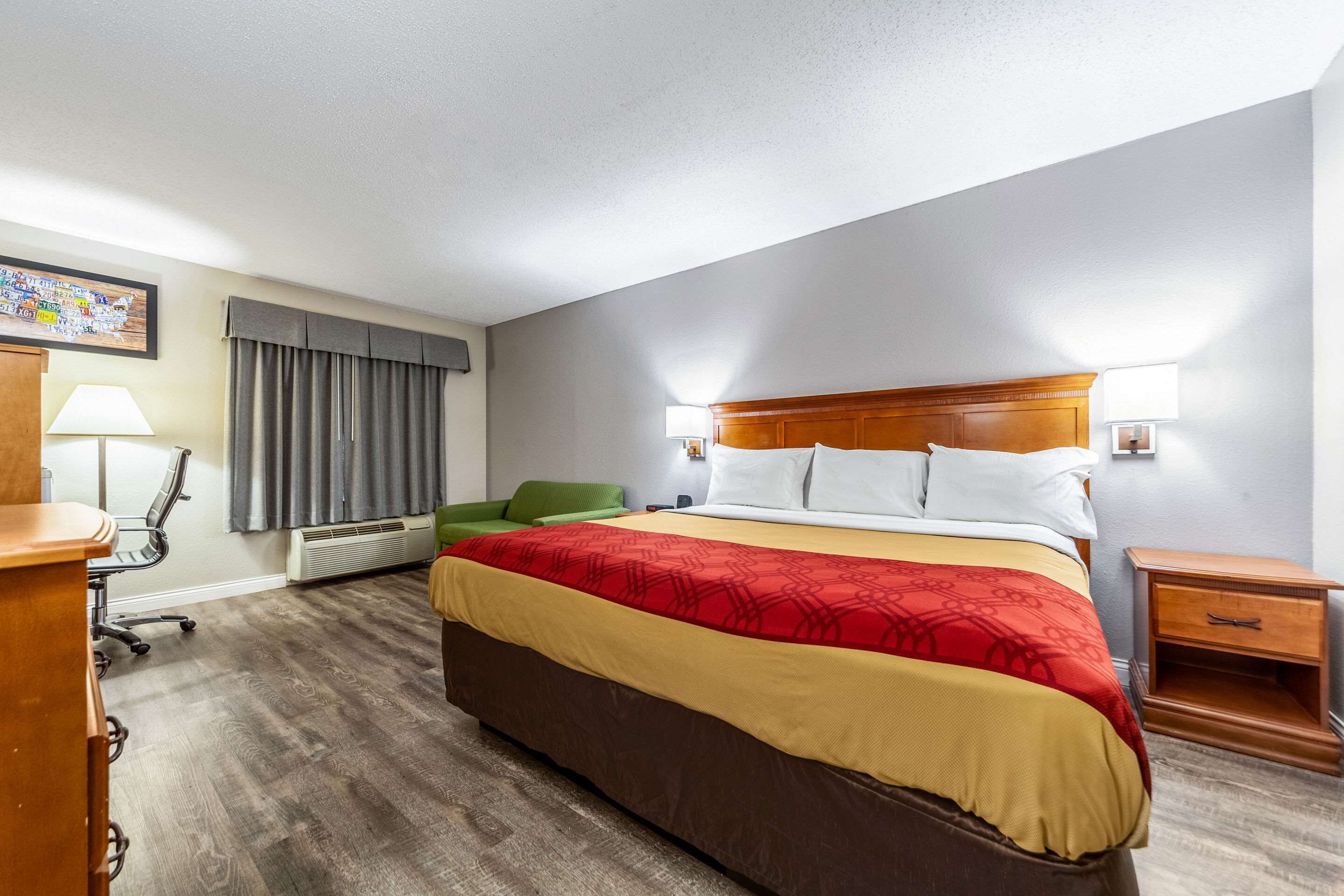 A hotel room with a large bed, a desk with a lamp, a green chair, and a small wooden nightstand.