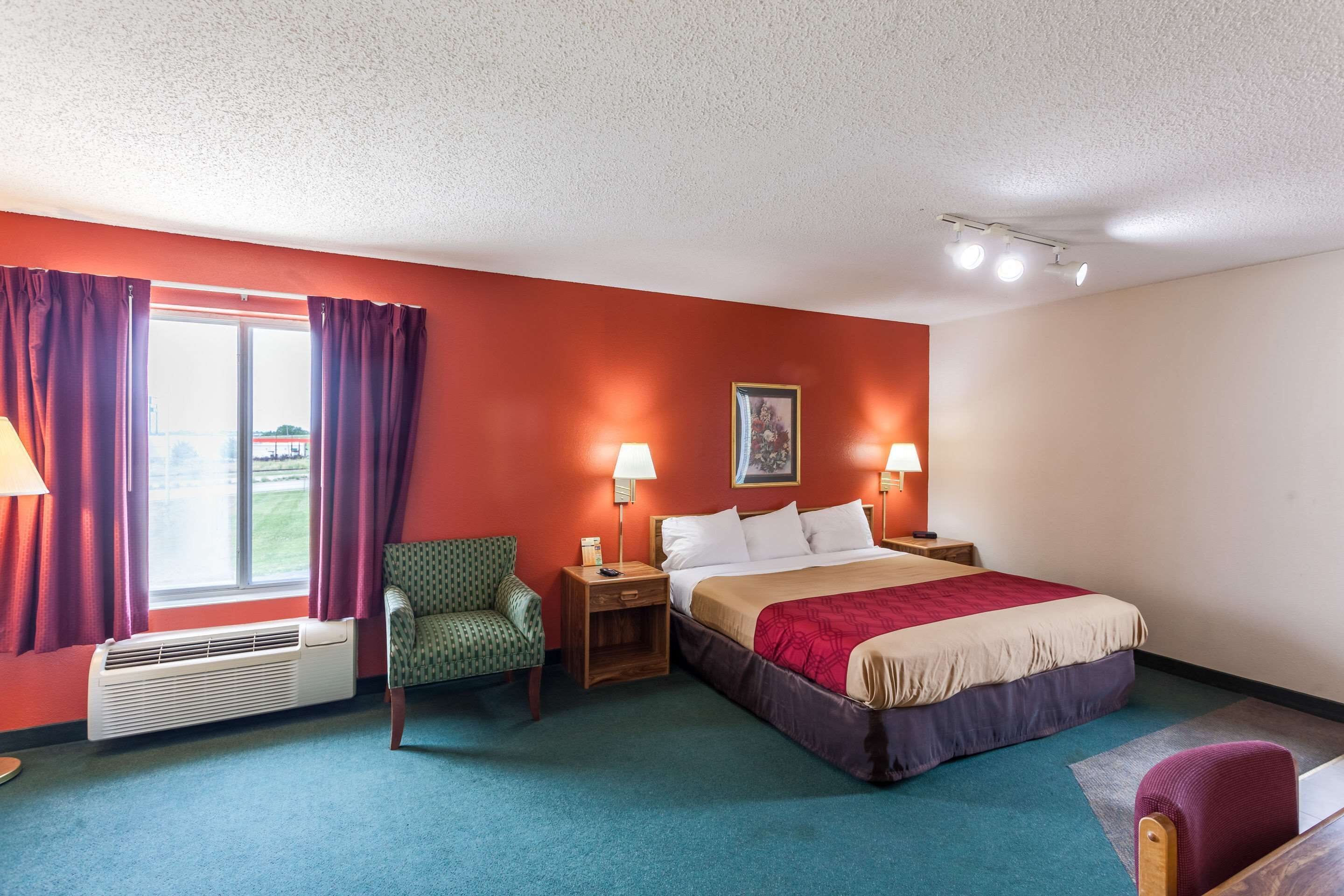 A hotel room with a large bed, two bedside tables with lamps, a chair, a window with curtains, and a radiator.