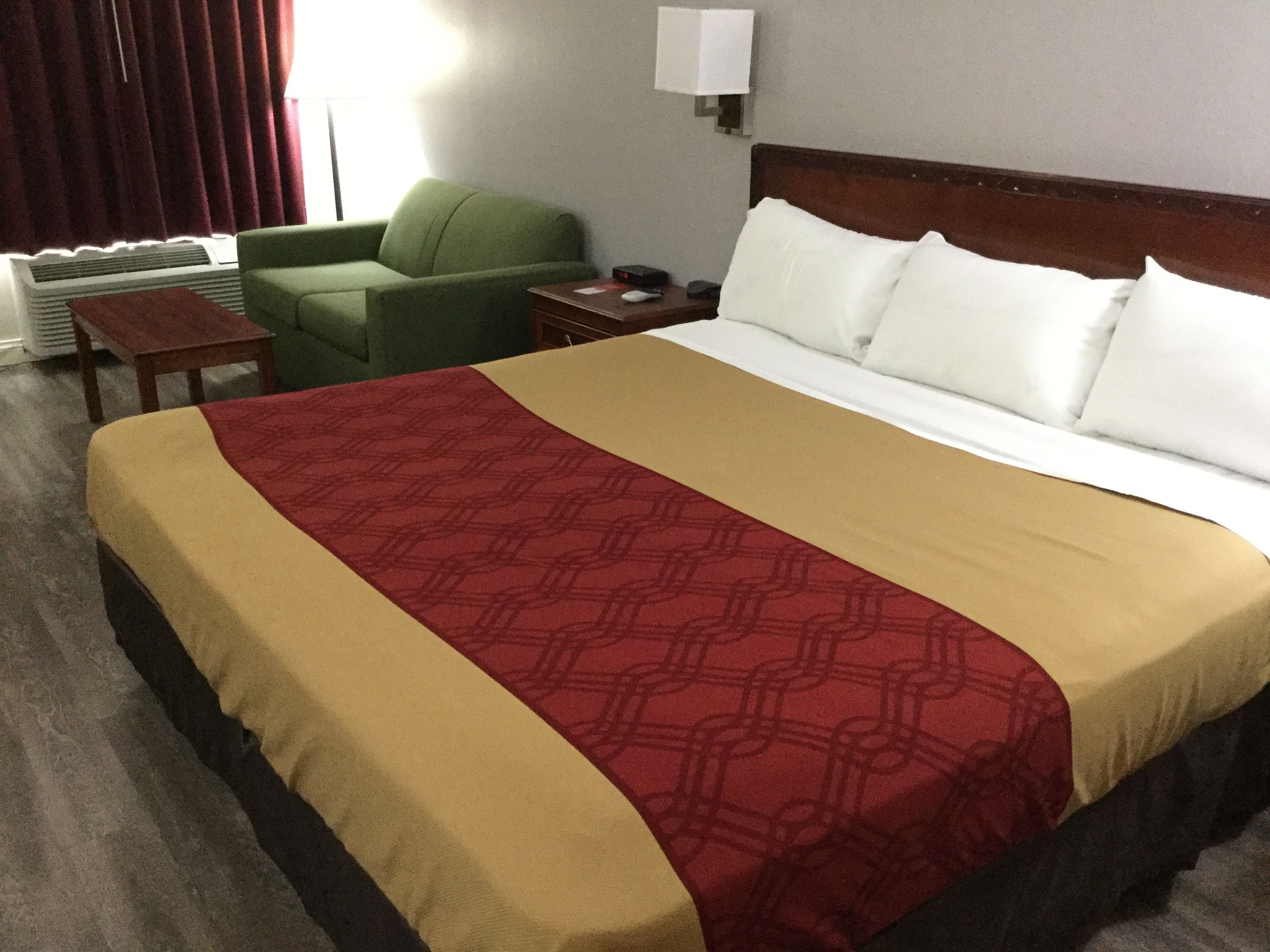 Quarto de hotel com uma cama grande, uma poltrona verde, uma mesinha e um criado-mudo com abajur.
