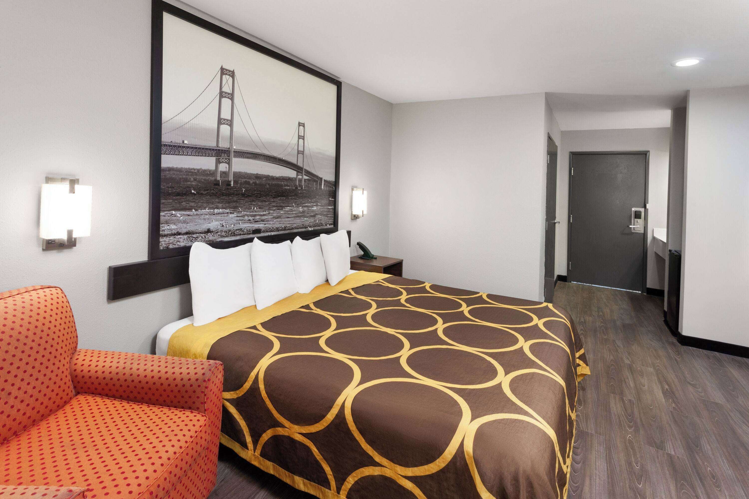 A hotel room with a bed, a chair, a painting of a bridge, and a door.