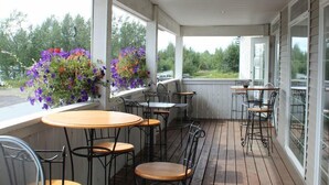 Een overdekt terras met houten vloer, metalen stoelen en tafels. Er staan bloembakken op de vensterbanken.