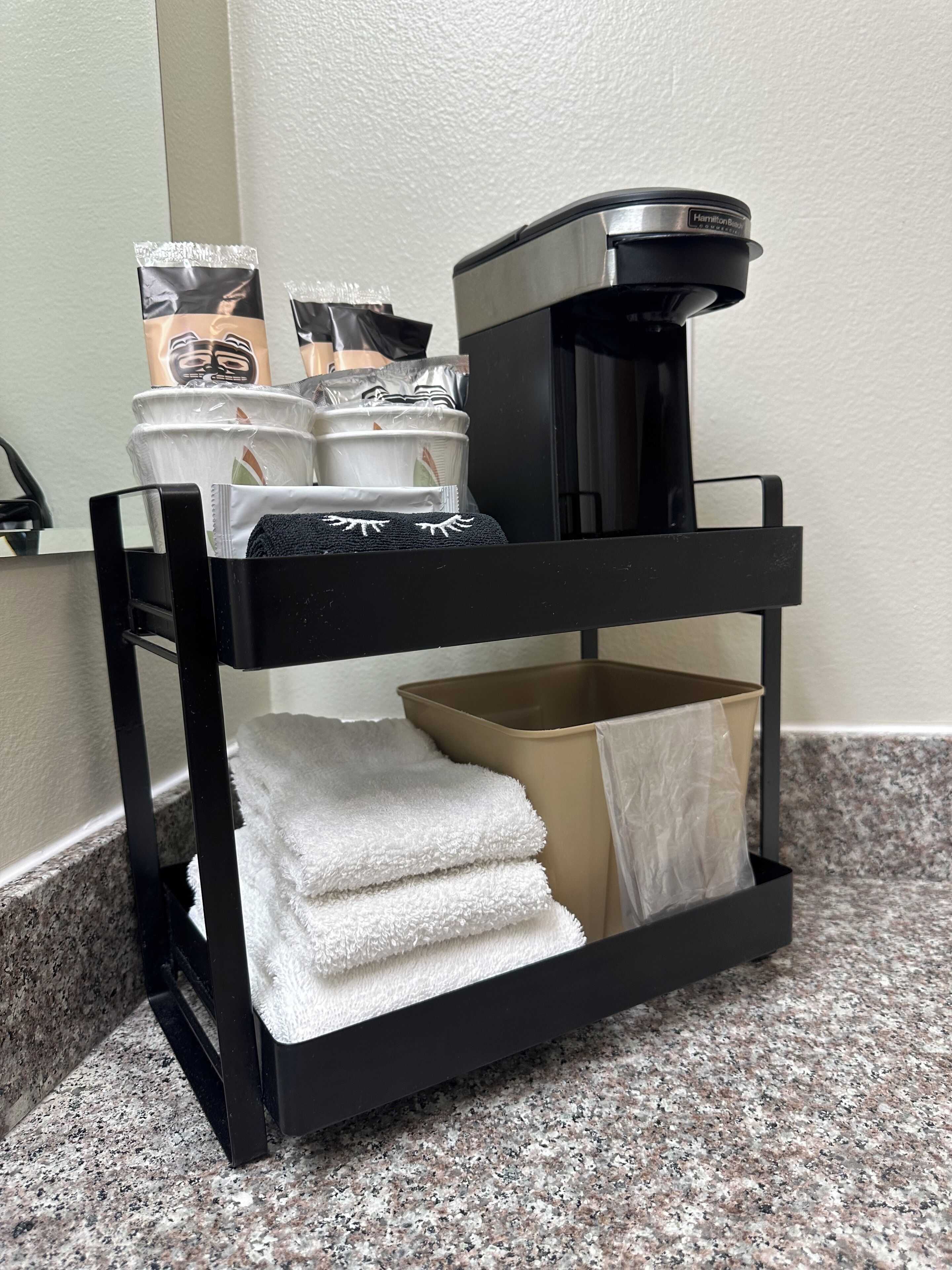 A black metal shelf with a coffee maker, stacked white towels, and a cardboard box.