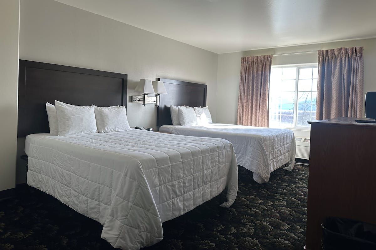 Quarto de hotel com duas camas, cabeceira escura, escrivaninha com computador e janela com cortinas.