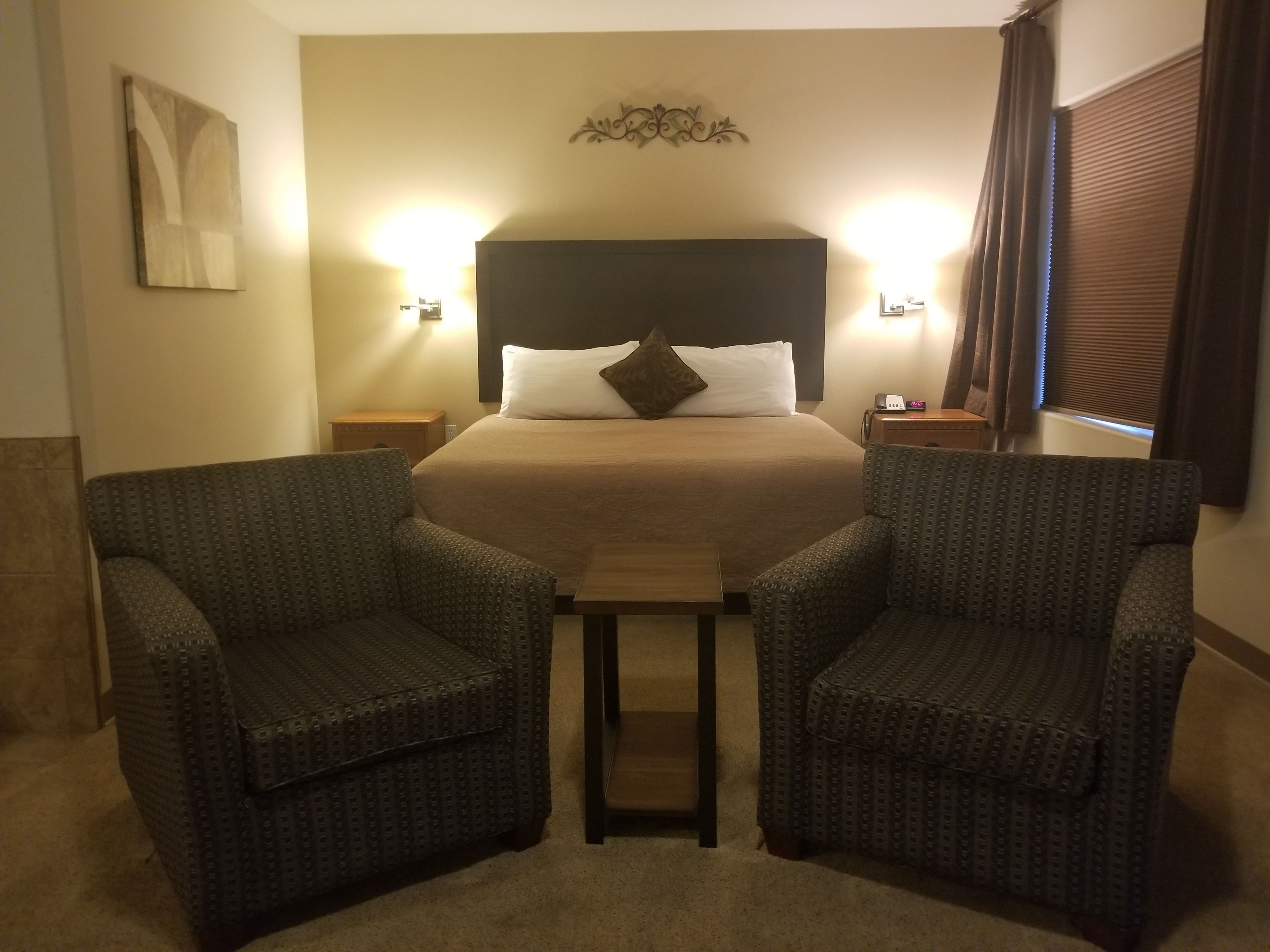 A hotel room with a large bed, two armchairs, a small table, and a nightstand with a lamp.