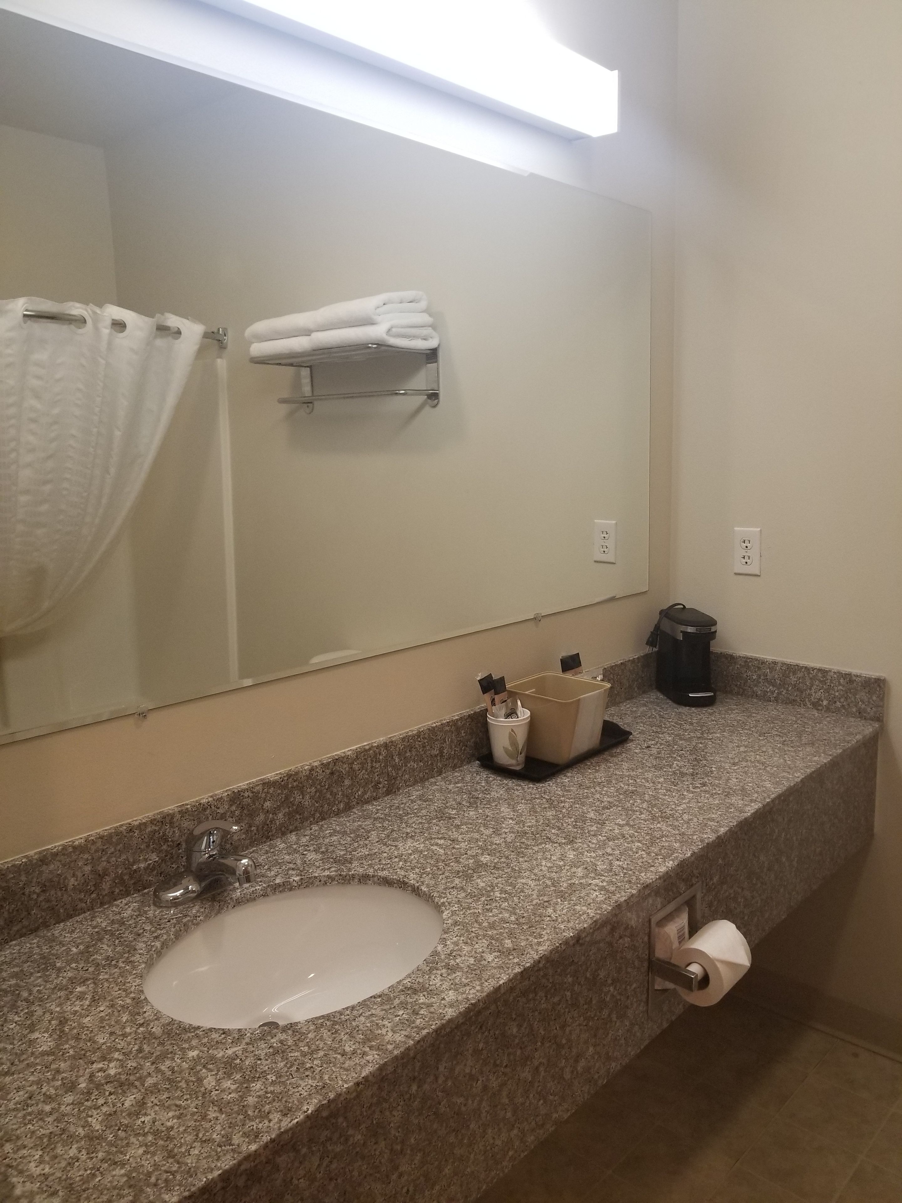 A bathroom with a granite countertop, a sink, a mirror, a towel rack, a trash can, and a coffee maker.