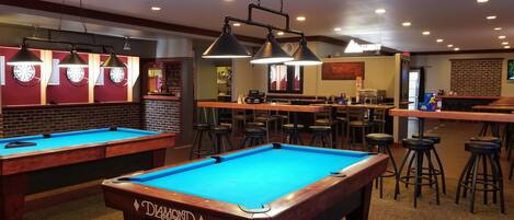 Una sala biliardo con due tavoli da biliardo, sgabelli da bar e illuminazione a sospensione.