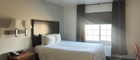 A hotel room with a bed, bedside table, lamp, and window with curtains.
