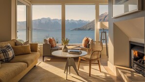 A cozy living room with a large window overlooking a lake and mountains. There is a sofa, a chair, a small table with books, and a fireplace. - Rosewood Matakauri (Closeburn)
